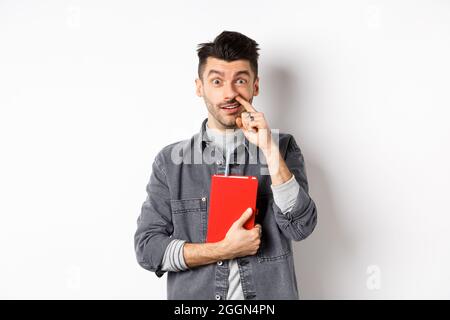 Junger Kerl Kommissionierung Nase und halten Buch, schauen dumm an der Kamera, stehen vor weißem Hintergrund Stockfoto