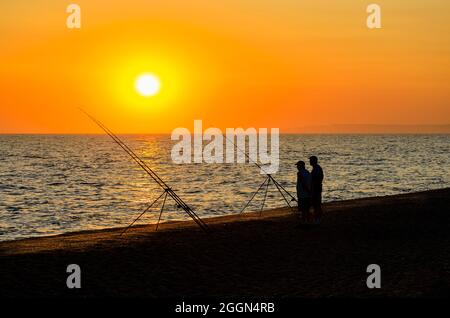 Angler fischen von festen Ruten am Strand in der Dämmerung, in West Bexington, einem Dorf im Südwesten Dorset, hinter dem Strand von Cesil in der Nähe von Bridport, bei Sonnenuntergang Stockfoto