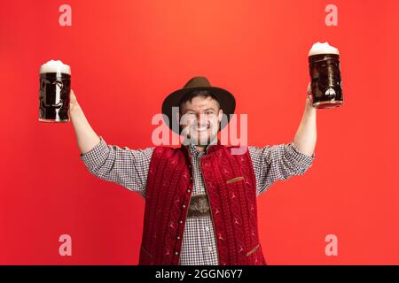 Fröhlicher lächelnder Mann in traditioneller österreichischer oder bayerischer Tracht mit zwei Tassen, Gläser dunkelschwarzen Bieres isoliert auf rotem Hintergrund Stockfoto