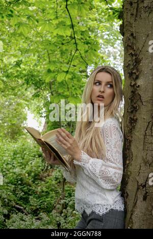 Schöne blonde Frau, die sich an einen Baum lehnt und ein Buch hält, sieht aus, als ob sie vom Lesen gestört wäre Stockfoto