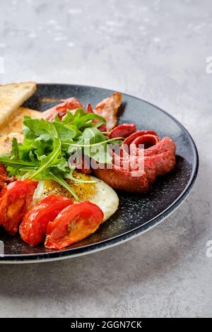 Nahaufnahme von Spiegeleiern mit Wurst, Speck, Tomaten und Bohnen auf einem Teller mit Toast Stockfoto