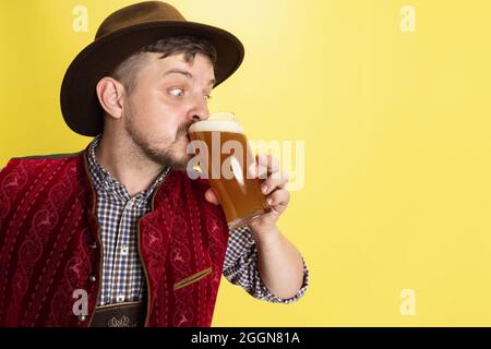 Nahaufnahme junger Mann in traditioneller bayerischer Tracht, der kaltes, geschäumtes Bier trinkt, Lagerbier, Ale isoliert auf gelbem Hintergrund. Nationale Küche Stockfoto