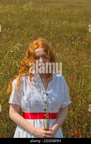 Rothaarige Mädchen trägt weißes Kleid in einem Feld von toten Mohnblumen mit einem Strauß von Mohnstielen Stockfoto