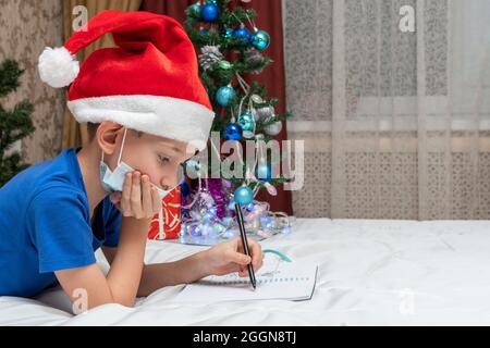 Neujahr und Weihnachtstraditionen während der Coronavirus-Sperre. Ein kleiner kaukasischer Junge in einer medizinischen Maske und einem roten Hut schreibt einen Brief an Santa AT Stockfoto