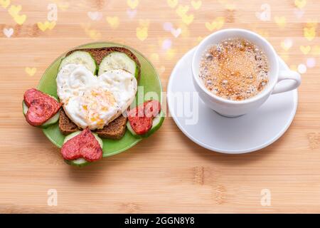 Kaffee in einer weißen Tasse und ein Sandwich mit herzförmigen Spiegeleiern, Würstchen und Gurken auf einem grünen Teller auf Holzboden mit Bokeh, Draufsicht Stockfoto