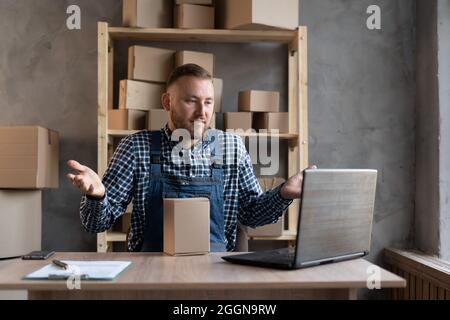 Ein männlicher Lieferdienstmitarbeiter sitzt in Overalls am Tisch und führt einen Videoanruf durch. Breitet seine Arme zu den Seiten aus, das Konzept eines verlorenen Produkts. Zoll Stockfoto