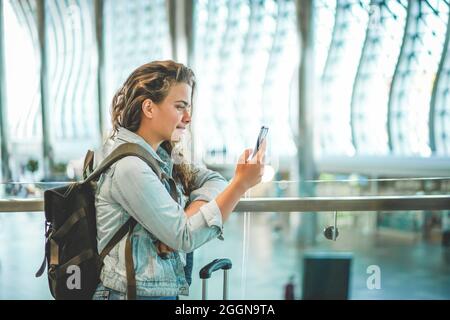 Eine wunderschöne kaukasische junge Frau, die am Flughafen ein Mobiltelefon benutzt, um Tickets online zu kaufen, ein Hotel zu buchen oder ein Auto zu mieten. Covid-Zeitreisen, Platz für Text Stockfoto