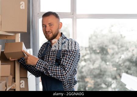 Der kaukasische bärtige Mann mit blauen Overalls legt eine flache Schachtel mit Waren in einem Lagerhaus auf ein Regal. Speicherplatz kopieren. Stockfoto