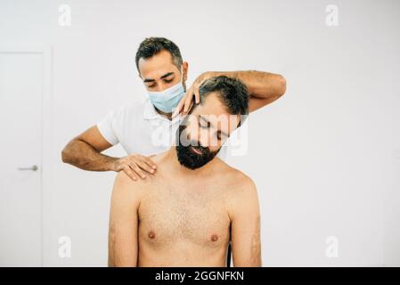 Männlicher Physiotherapeut mit Maske arbeitet an der Genesung eines Patienten in der Physiotherapie-Klinik. Der schwarzbärtige Patient sitzt und wird behandelt Stockfoto