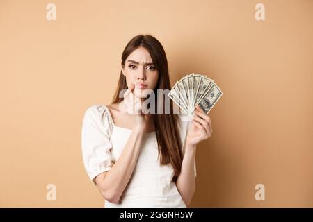 Nachdenkliche ernsthafte Mädchen hält Geld und Denken, Blick nachdenklich an der Kamera und runzelte die Stirn, entscheiden, was mit Dollarnoten kaufen, beige Hintergrund Stockfoto