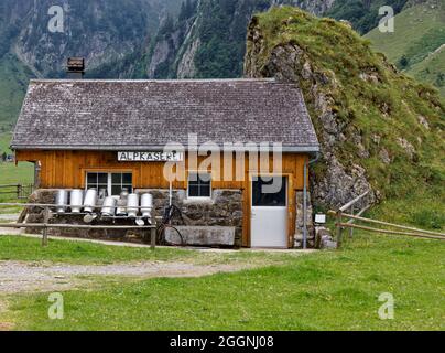 schweizer Almkäserei im alten Stil gebaut neben einer Felswand leere Milchdosen vor dem Haus, deutsches Almkäsereizeichen, Text transl Stockfoto