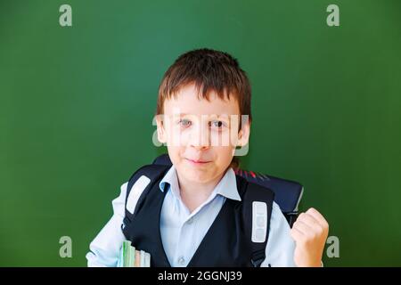 Lustige Schuljunge Grimassen in der Nähe der grünen Schultafel im Klassenzimmer. Grundschulkind mit Tasche und Büchern. Zurück zur Schule. Stockfoto