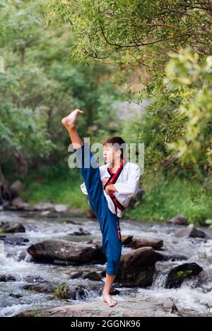 Das japanische Kind praktiziert Taekwondo mit einem Kimono der Kampfkunst in der Natur. Schwarzes Karate-Gürtel-Training mit hochgefahrtem Bein. Stockfoto