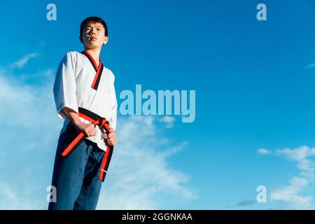 Porträt eines jungen Japaners in seinem Taekwondo-Kimono, der den schwarzen Gürtel seines Kampfkünste-Kimonos hält. Weißer Kimono und blaue Karate-Hose Stockfoto