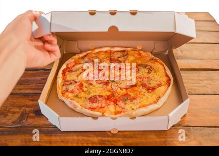 Die Hand des Typen öffnet eine Pappschachtel Pizza vor dem Hintergrund eines Holztisches. Köstliches Fast Food. Stockfoto