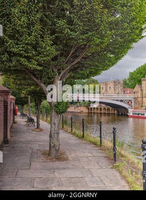 Ein Pfad mit Bäumen und Bänken verläuft entlang eines Flusses. Eine alte Brücke und historische Gebäude sind im Hintergrund mit einem Touristenboot in der Nähe. Stockfoto