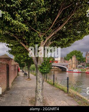 Ein Pfad mit Bäumen und Bänken verläuft entlang eines Flusses. Eine alte Brücke und historische Gebäude sind im Hintergrund mit einem Touristenboot in der Nähe. Stockfoto