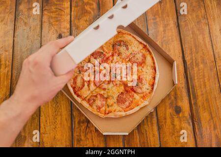 Die Hand des Typen öffnet eine Pappschachtel Pizza vor dem Hintergrund eines Holztisches. Köstliches Fast Food. Stockfoto