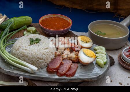 Rotes Schweinefleisch mit Reis (Kao Moo Dang): Rotes Grillpork im thailändischen Stil, Entenei und knuspriger Schweinebauch, geschnittene Gurke mit roter Soße und Brühe. Stockfoto