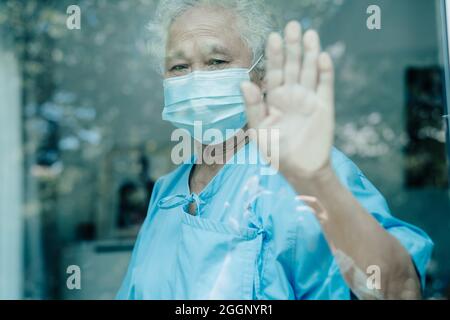 Asiatische alte ältere Patientin mit Maske, die während einer Covid19-Coronavirus-Pandemie aus dem Fenster schaut, um sich in Quarantäne zu begeben, bleibt zu Hause. Stockfoto