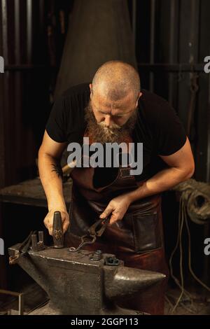 Bärtiger Mann, Schmied, der das geschmolzene Metall mit Funkenfeuerwerk in Schmiede auf dem Amboss manuell schmiedet. Konzept der Arbeit, Retro-Berufe, Familie Stockfoto