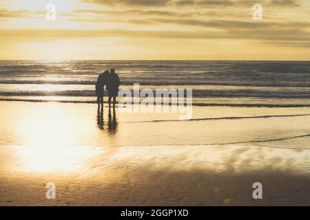 Ein im Ruhestand gealtertes Paar steht weg von der Kamera und blickt auf das Meer, während sie sich im Sonnenuntergang zusammen entspannen Stockfoto