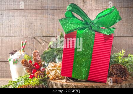 Große rote Weihnachts-Geschenkbox mit glitzerndem, festlich grünem Band, auf traditionellem Holzhintergrund mit Weihnachtsdekor und Lebkuchenkuchen, Kopierraum Stockfoto