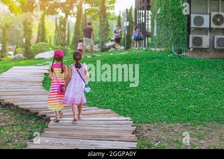 Rückansicht von asiatischen kleinen Mädchen im Kleid, die Seite an Seite auf dem Weg durch den grünen Garten gehen.zwei Freundinnen, die zusammen im Park gehen. Frohe Freunde Stockfoto