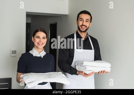 Lächelnde Zimmermädchen hielten weiße Bettwäsche und Handtücher im Hotelzimmer Stockfoto