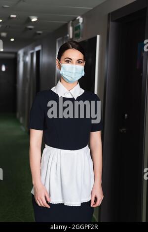 Junge Zimmermädchen in medizinischer Maske und Uniform, die auf die Kamera im Flur des Hotels schaut Stockfoto