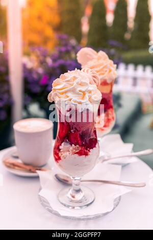 Erdbeereis-Dessert mit Schlagsahne und zerdrückter Baiser, serviert in einer Glastasse. Eine Tasse Kaffee auf dem Couchtisch. Stockfoto