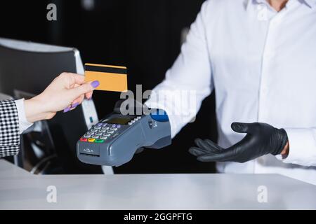 Abgeschnittene Ansicht der Empfangsdame in Latexhandschuhen, die das Zahlungsterminal mit Kreditkarte in der Nähe des Touristen halten Stockfoto