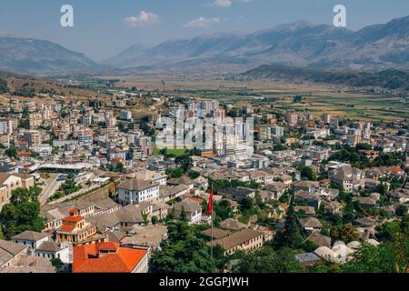 Stadtbild Gjirokastra – traditionelle albanische weiße Häuser mit steinernen grauen Dächern und modernen Gebäuden. Sommerlandschaft mit üppigem Laub, blauem Himmel und Stockfoto