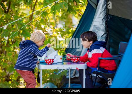 Glückliche Kinder, Frühstück am Morgen vor einem Zelt auf einem Campingplatz im Wald in Norwegen, sonnig am frühen Morgen Stockfoto