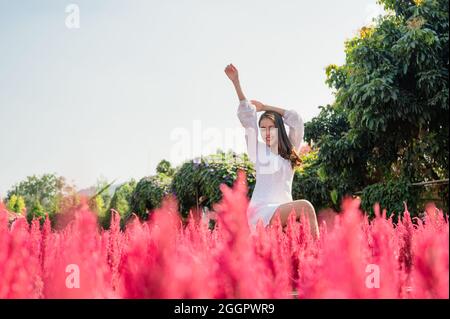 Fröhliche schöne junge asiatische Frau, die mit aufhebenden Armen in einer im Garten blühenden Kakerlakenblüte sitzt Stockfoto