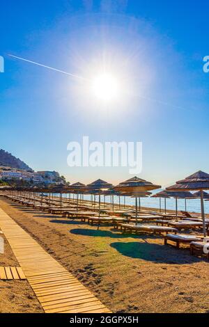 Chemtrail Überqueren Sie die Sonne über Liegestühlen und Sonnenschirmen und den blauen Himmel am Strand auf der Insel Kos in Griechenland. Stockfoto