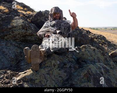Foto eines gemieteten Scharfschützen in Tarnung während der Ruhe. Er sitzt auf den Felsen und zeigt seine Daumen nach oben. Konzept einer professionellen Armee bei bewaffneten Konflikten Stockfoto