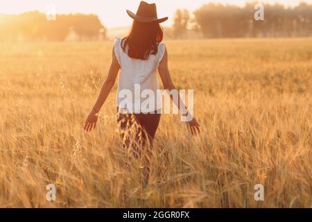 Frau Bäuerin im Cowboy Hut, die bei Sonnenuntergang mit Händen auf Ohren beim landwirtschaftlichen Weizenfeld läuft. Stockfoto