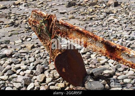 Ein Bild eines alten, rostigen Spatenankers, der an einem steinigen Strand aufgegeben wurde, dessen Spitze in die Steine geklebt war Stockfoto