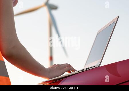 Nahaufnahme Frau Hände arbeiten auf dem Laptop auf der Motorhaube eines Autos vor dem Hintergrund einer Windkraftanlage. Wartung der Windmühle zur Erzeugung von Stockfoto