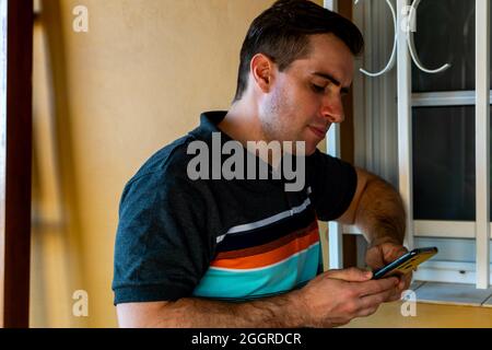 Der junge kaukasische Mann benutzte sein Mobiltelefon, während er sich auf die Fensterbank lehnte Stockfoto