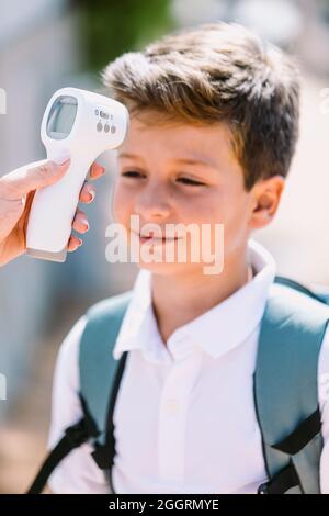 Der Lehrer nimmt die Temperatur eines Kindes, wenn er mit einem elektronischen Thermometer in die Schule geht. Covid-19 und Virenpandemic-Konzept Stockfoto