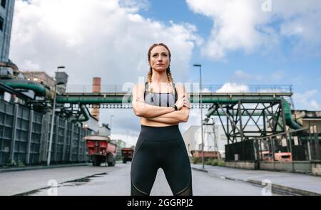 Sportliche Frau mit gekreuzten Armen, die vor einer Fabrik posiert Stockfoto
