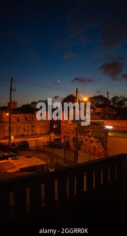 Orangefarbene Stadtlichter, Fenster, Straßen unter blauem und purpurfarbenem Sonnenuntergangshimmel mit Halbmond Stockfoto