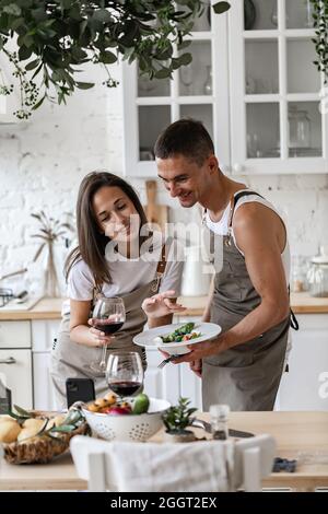 Glückliches Paar, das Videoanruf mit dem Smartphone macht, während es das Abendessen in der Küche zu Hause zubereitet fröhliche kaukasische Menschen winken ihre Hände und lächeln Stockfoto