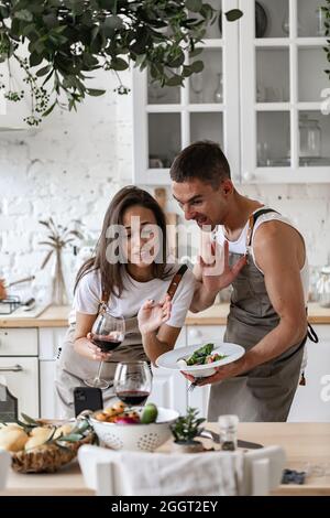 Glückliches Paar, das Videoanruf mit dem Smartphone macht, während es das Abendessen in der Küche zu Hause zubereitet fröhliche kaukasische Menschen winken ihre Hände und lächeln Stockfoto