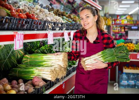 Junge lächelnde Verkäuferin legte Lauch auf den Tresen aus Stockfoto