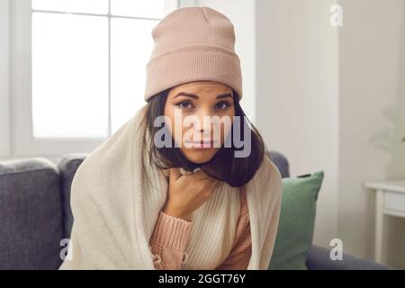 Kranke Frau in einem warmen Pullover und Hut sitzt auf dem Sofa zu Hause in einem karierten gewickelt. Stockfoto