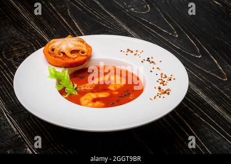 Schüssel Tomatensuppe mit Garnelen mit einem kleinen Oktopus auf einer orangefarbenen Scheibe auf schwarzem Holzgrund gebacken, Draufsicht Stockfoto