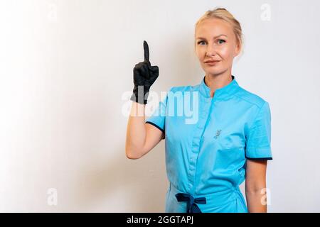 Medizin und Schönheit. Eine junge Frau in Uniform und Gummihandschuhen zeigt ihren Finger nach oben. Weißer Hintergrund. Speicherplatz kopieren. Medizinisches Personal, cosmetolo Stockfoto